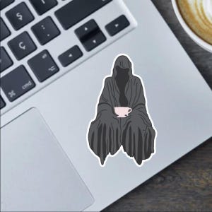 Könnte beinhalten: Ein Aufkleber mit einer dunklen Gestalt in einer Kapuzenrobe, die eine rosa Teetasse hält. Die Gestalt sitzt, wobei die Robe sie umhüllt. Der Aufkleber befindet sich auf einem Laptop, mit einer sichtbaren Tastatur.