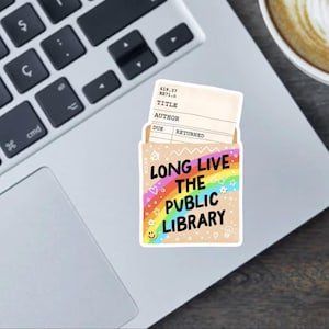 May include: A sticker with a library theme, featuring a rainbow and the text "LONG LIVE THE PUBLIC LIBRARY." The sticker includes a library card design and is placed on a laptop. The sticker has a light brown background.