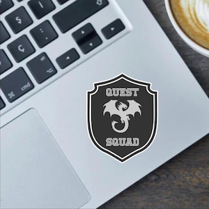 May include: A black and white sticker with a shield design. The shield has a gray dragon inside and the words "QUEST SQUAD" written on it.