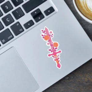 May include: A pink sticker featuring a sword design adorned with orange roses, pink flowers, and green leaves. The sword has a pink handle and a crossguard. The sticker is on a laptop.