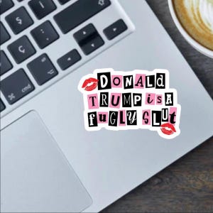 Op de afbeelding: Een witte sticker met de tekst "DONALD TRUMP is a fugly slut" in collage-stijl, met roze en zwarte blokletters en rode lippenstiftkusjes. De sticker zit op een laptop.
