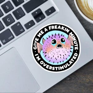 Puede incluir: Una pegatina redonda con un diseño de pez globo de dibujos animados con una expresión angustiada, lágrimas y una combinación de colores rosa, morado y naranja. La pegatina tiene el texto "I'M OVERSTIMULATED. GIVE ME A FREAKIN MINUTE."