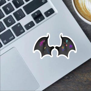 Könnte beinhalten: Schwarze Flügel mit bunten Farbflecken auf einem weißen Laptop.
