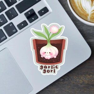 May include: A sticker featuring a cartoon garlic bulb with a smiling face, sprouting green leaves and a pink flower, in a brown pot. The text "garlic gori" is below the pot.