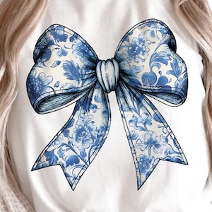 May include: A white t-shirt with a blue and white floral bow design. The bow is large and centered on the shirt. The shirt is a casual style with a crew neck and short sleeves.