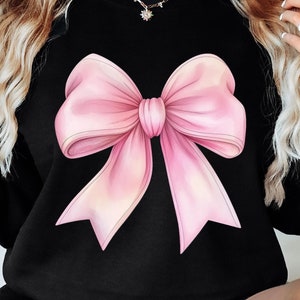 May include: A black sweatshirt with a large pink bow graphic on the front. The bow is a light pink color with a darker pink center.
