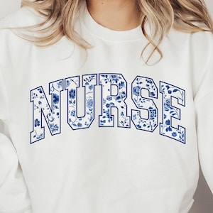 May include: White sweatshirt with the word "NURSE" in large, blue floral print letters across the chest. The design features a classic collegiate font with a floral pattern within each letter. The bottom of the image includes the text "SVG PNG" and "DIGITAL DOWNLOAD".