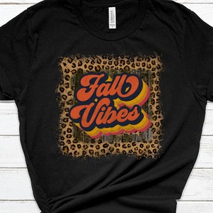 May include: Black t-shirt with a distressed leopard print frame around the text "Fall Vibes" in a retro font with orange, yellow, and blue colors.