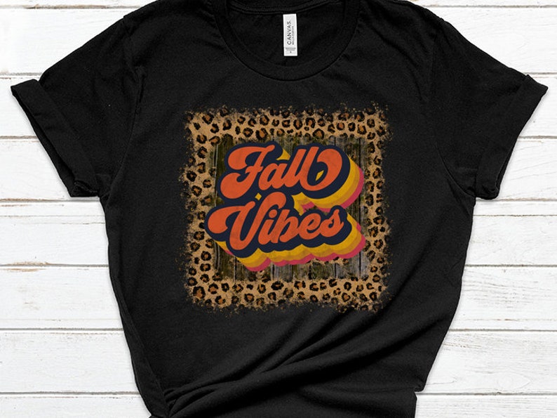 May include: Black t-shirt with a distressed leopard print frame around the text "Fall Vibes" in a retro font with orange, yellow, and blue colors.