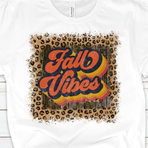 May include: White t-shirt with a distressed graphic design featuring the text "Fall Vibes" in a retro font with orange, yellow, and blue colors. The graphic is surrounded by a leopard print pattern.