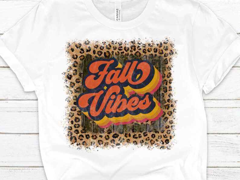 May include: White t-shirt with a distressed graphic design featuring the text "Fall Vibes" in a retro font with orange, yellow, and blue colors. The graphic is surrounded by a leopard print pattern.