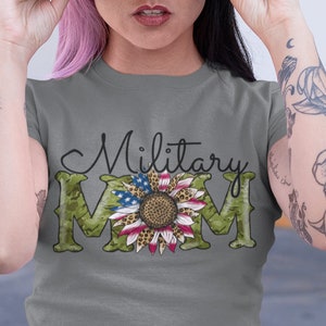 Może przedstawiać: Szary T-shirt z napisem "Military Mom" czarnymi literami. Litera "M" to wz&oacute;r kamuflażu z słonecznikiem w środku. Słonecznik ma czerwone, białe i niebieskie płatki i leopardzi wz&oacute;r w środku.