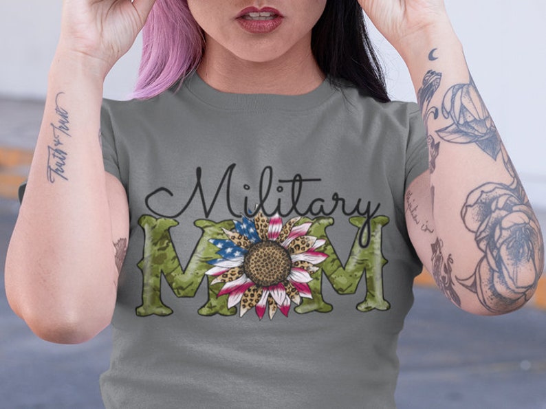 Może przedstawiać: Szary T-shirt z napisem "Military Mom" czarnymi literami. Litera "M" to wz&oacute;r kamuflażu z słonecznikiem w środku. Słonecznik ma czerwone, białe i niebieskie płatki i leopardzi wz&oacute;r w środku.