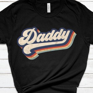May include: Black t-shirt with a retro-style graphic that says "Daddy" in a colorful, faded rainbow design.