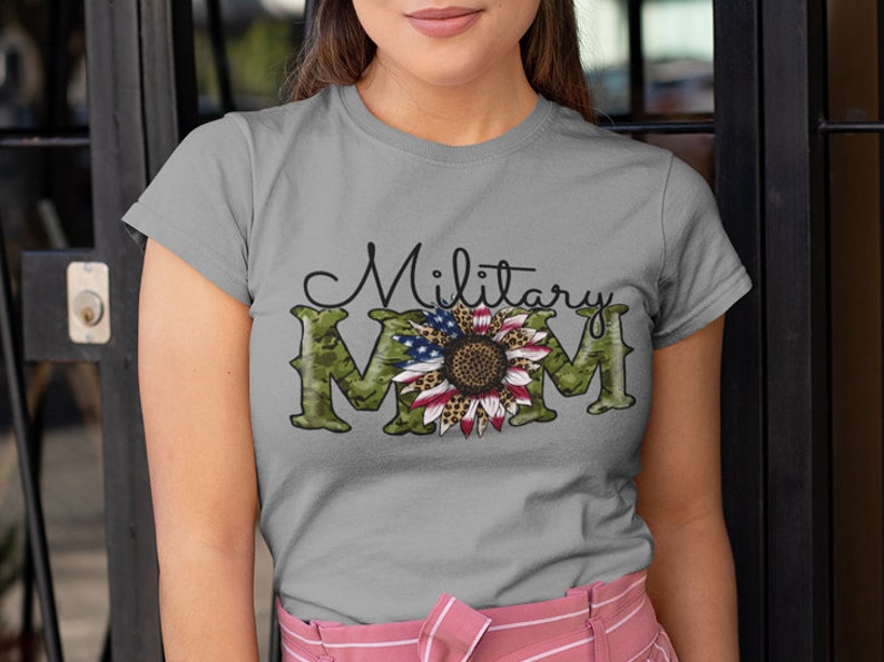 Może przedstawiać: Szary T-shirt z zielonym nadrukiem kamuflażu i słonecznikiem z wzorem amerykańskiej flagi. Na koszulce nadrukowany jest tekst "Military Mom".
