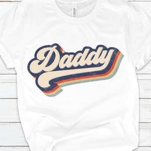 May include: White t-shirt with a retro-style graphic that says "Daddy" in a colorful rainbow design.