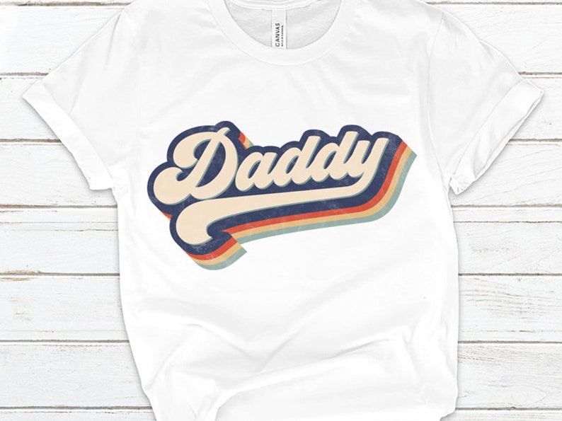May include: White t-shirt with a retro-style graphic that says "Daddy" in a colorful rainbow design.