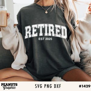 May include: A dark gray t-shirt with the text "Retired Est 2025" in white lettering. The shirt is being worn by a person.