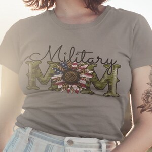 Może przedstawiać: Szary T-shirt z napisem "Military Mom" w czarnym kolorze. Litera "M" to wz&oacute;r kamuflażu z słonecznikiem w środku. Słonecznik ma czerwone, białe i niebieskie płatki, przypominające polską flagę.