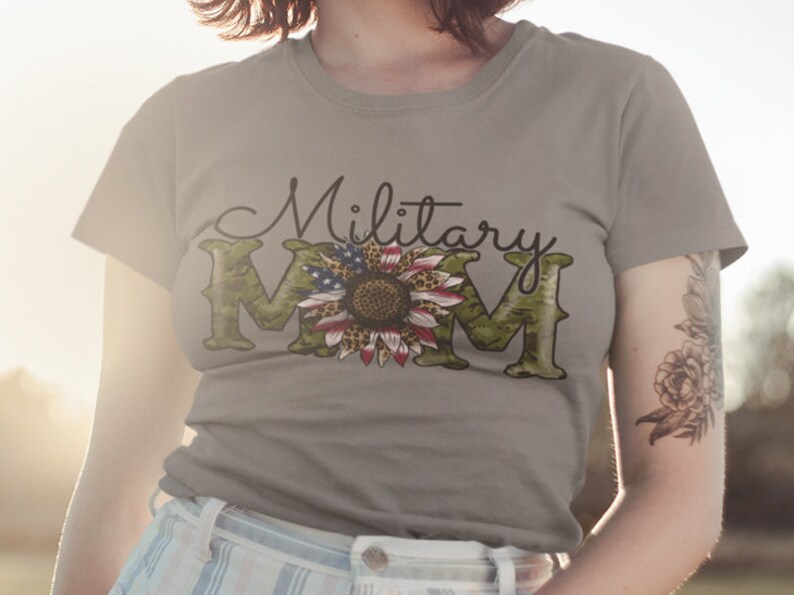 Może przedstawiać: Szary T-shirt z napisem "Military Mom" w czarnym kolorze. Litera "M" to wz&oacute;r kamuflażu z słonecznikiem w środku. Słonecznik ma czerwone, białe i niebieskie płatki, przypominające polską flagę.