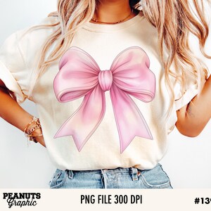 May include: A white t-shirt with a large pink bow graphic. The bow is drawn in a watercolor style with soft edges.