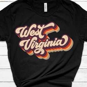 K&ouml;nnte beinhalten: Schwarzes T-Shirt mit einer Retro-Grafik, die "West Virginia" in bunten, &uuml;bereinanderliegenden Buchstaben zeigt.