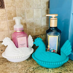 May include: Two decorative soap dispensers in chicken-shaped holders. One dispenser is pink with "Champagne Toast" text, the other is dark blue with "Coconut Angel Cake" text. The holders are white and turquoise.