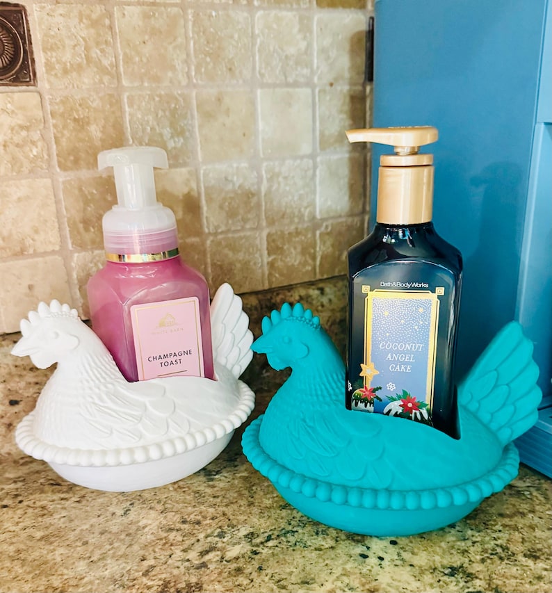 May include: Two soap dispensers in chicken-shaped holders. One dispenser is pink with "Champagne Toast" text, the other is dark blue with "Coconut Angel Cake" text. The holders are white and turquoise.
