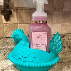 May include: A teal chicken-shaped soap dispenser holder. It holds a pink soap bottle labelled "Champagne Toast" from White Barn. The dispenser has a white pump and is set against a neutral-toned tile background.