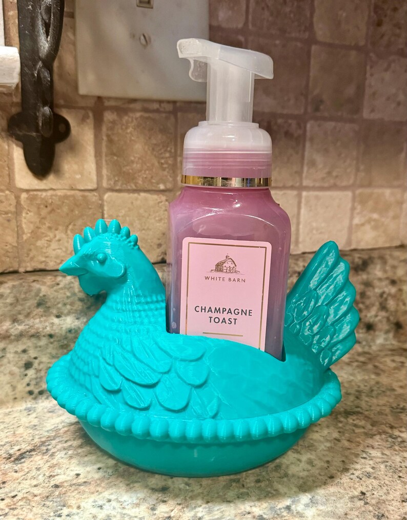 May include: A teal chicken-shaped soap dispenser holder. It holds a pink soap bottle labelled "Champagne Toast" from White Barn. The dispenser has a white pump and is set against a neutral-toned tile background.