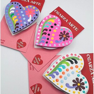 May include: Three wooden heart-shaped ornaments with colorful painted designs. Each heart features a flower and swirls. The hearts are displayed on pink cards with the text "CGARZA ARTE" and a website address.