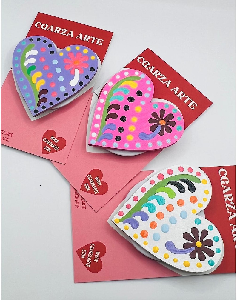 May include: Three wooden heart-shaped ornaments with colorful painted designs. Each heart features a flower and swirls. The hearts are displayed on pink cards with the text "CGARZA ARTE" and a website address.