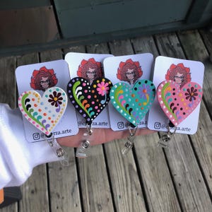 May include: Four wooden heart-shaped badge holders with colorful floral designs and polka dots. Each heart is attached to a retractable badge reel. The hearts are painted in black, white, pink, and turquoise.