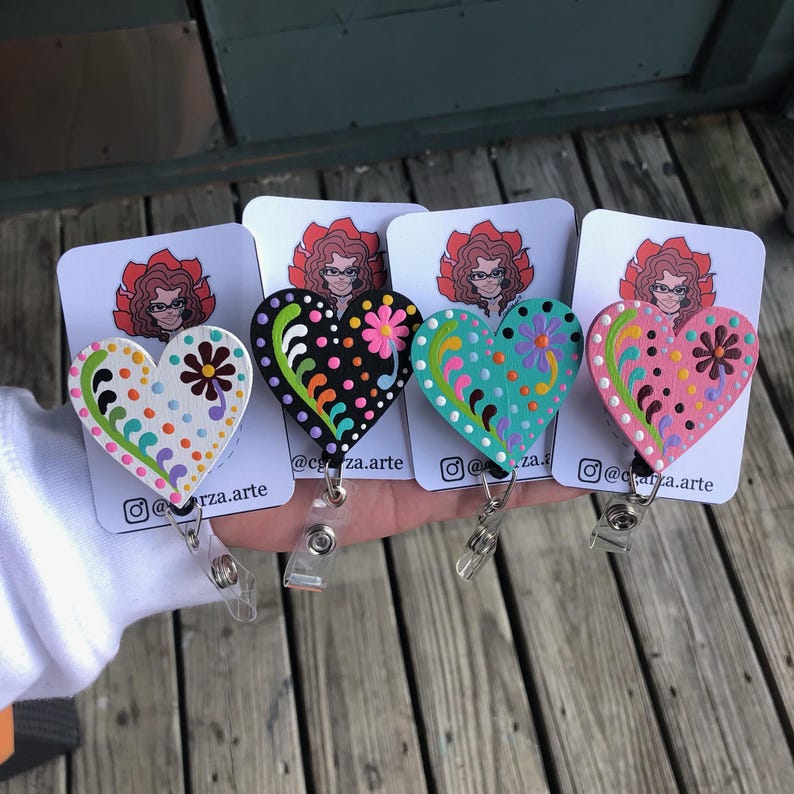 May include: Four wooden heart-shaped badge holders with colorful floral designs and polka dots. Each heart is attached to a retractable badge reel. The hearts are painted in black, white, pink, and turquoise.