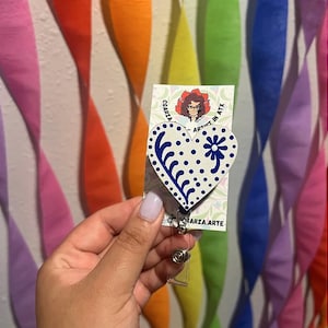 May include: A white heart-shaped badge holder with a blue and white floral design. The badge holder is attached to a clear plastic retractable badge reel. The badge holder is on a card with the text "CGARZA - ARTIST IN ATX" and "CGARZA.ARTE".