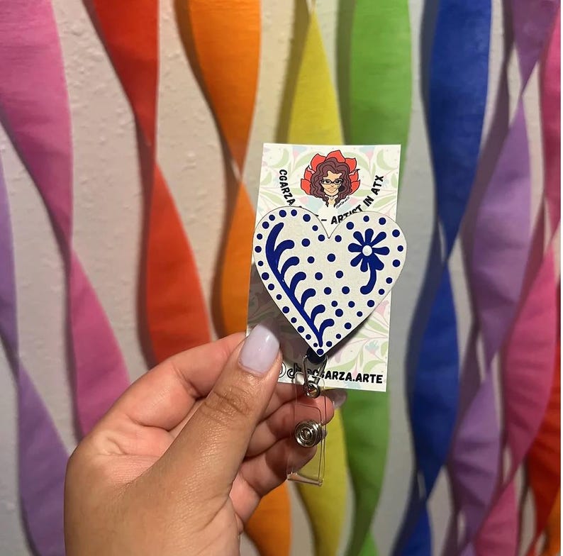 May include: A white heart-shaped badge holder with a blue and white floral design. The badge holder is attached to a clear plastic retractable badge reel. The badge holder is on a card with the text "CGARZA - ARTIST IN ATX" and "CGARZA.ARTE".