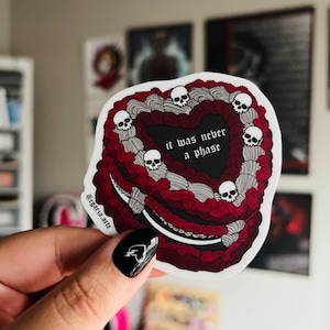 May include: A heart-shaped sticker featuring a dark red and grey piped frosting design with five small white skulls nestled in the icing. The center of the heart is black with the white text "it was never a phase" and the handle @cgarza.arte.