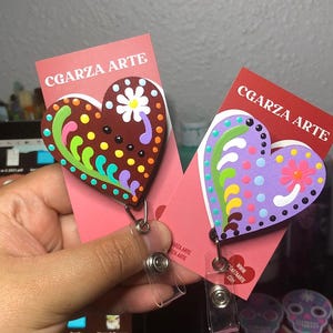 May include: Two wooden heart-shaped badge holders with colorful floral designs. The hearts are painted with purple, green, yellow, pink, and brown accents. The text "CGARZA ARTE" is printed on the cards.