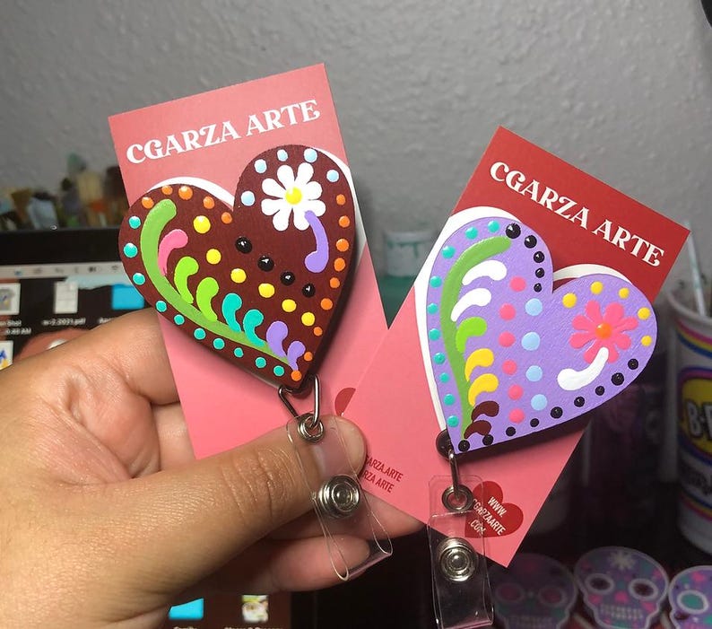 May include: Two wooden heart-shaped badge holders with colorful floral designs. The hearts are painted with purple, green, yellow, pink, and brown accents. The text "CGARZA ARTE" is printed on the cards.