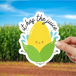 May include: A yellow corn on the cob with a smiling face and green leaves. The text "it has the juice" is written around the corn.