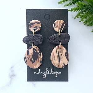 May include: A pair of dangle earrings with a brown, black, and beige marbled design. The earrings are made of polymer clay and have gold-toned hardware. The earrings are on a black card with the text "midnightclayco".