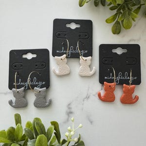 May include: Three pairs of cat-shaped dangle earrings on black cards with the text "midnightclayco". The earrings are in gray, white, and orange.