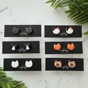 May include: Six pairs of cat-shaped polymer clay earrings on black cards. The earrings are in various colors, including black, white, orange, and brown. The cards have the text "midnightclayco" printed on them.