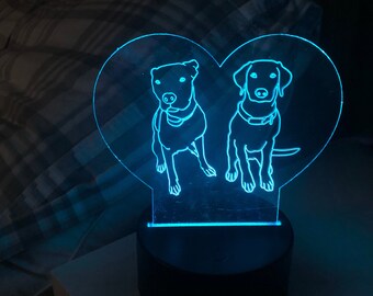 May include: A blue LED night light shaped like a heart with two dog silhouettes inside. The night light is on a white bedside table.