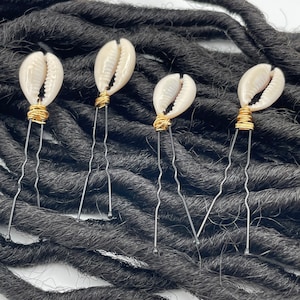 Puede incluir: Cinco horquillas para el pelo de concha de cauri con detalles de alambre dorado. Las horquillas son negras y están sobre un lecho de rastas negras.