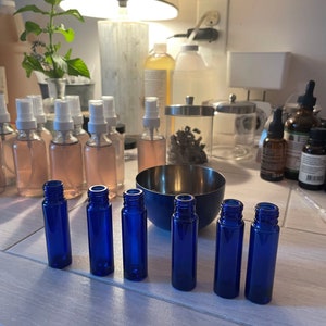 Puede incluir: Una colección de botellas de vidrio azul para aceites esenciales y botellas con atomizador. Las botellas están dispuestas sobre una superficie clara, con un cuenco de metal y otras botellas en el fondo. Las botellas están vacías y listas para ser llenadas.