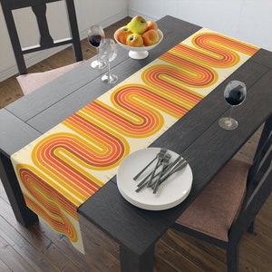 May include: A yellow, orange, and brown table runner with a retro geometric pattern of curved lines. The runner is on a dark wood table with a bowl of apples, two wine glasses, and a plate with silverware.