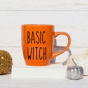 May include: Orange ceramic mug with black text that reads "BASIC WITCH". The mug is sitting on a white surface with decorative pumpkins.