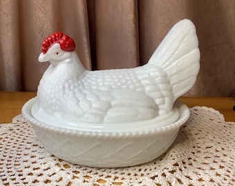 May include: White milk glass chicken shaped dish with a red comb. The chicken is sitting in a nest with a white and gray woven pattern.