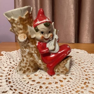 May include: A ceramic figurine of an elf playing a musical instrument, sitting on a tree stump. The elf is wearing a red hat and red clothing. The tree stump is brown and has a white glaze.